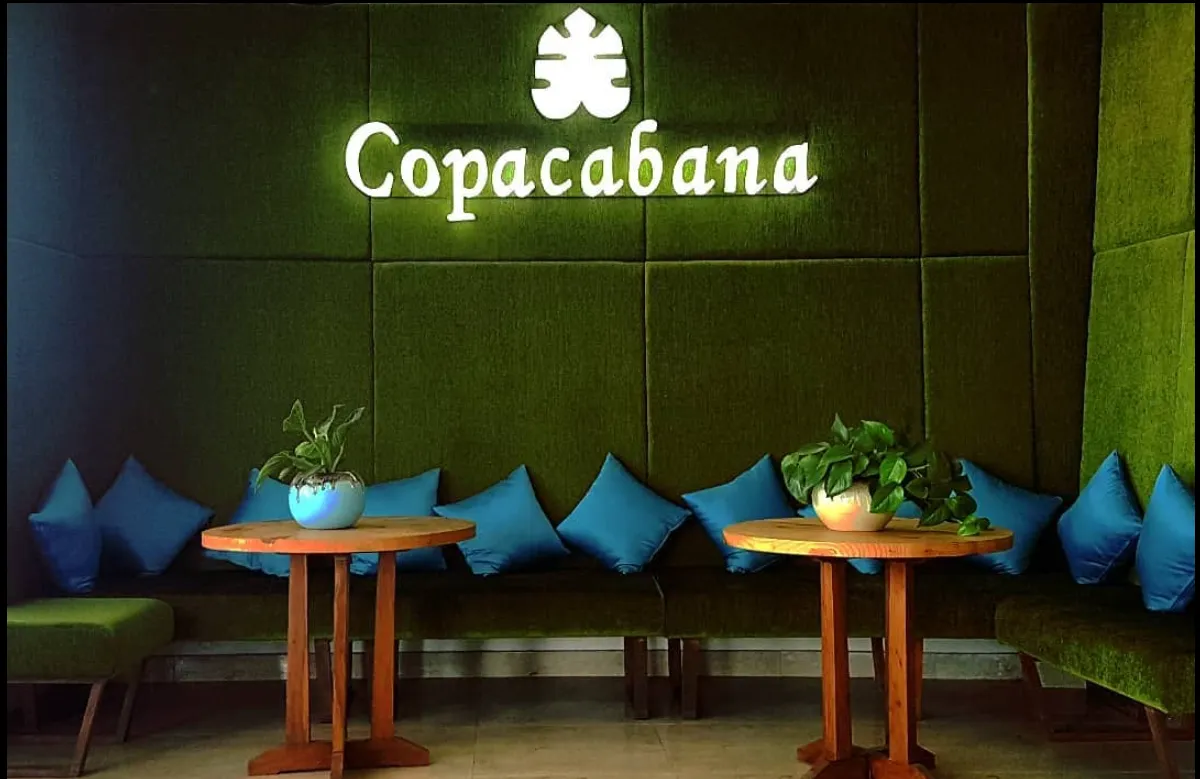 The illuminated Copacabana clover logo on a deep teal wall above turquoise banquette seating