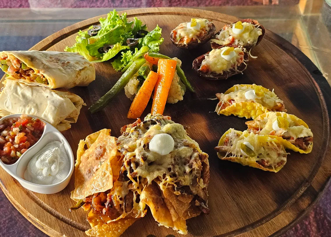 A wooden platter of burritos, loaded nachos, tacos and potato skins with salsa and sour cream