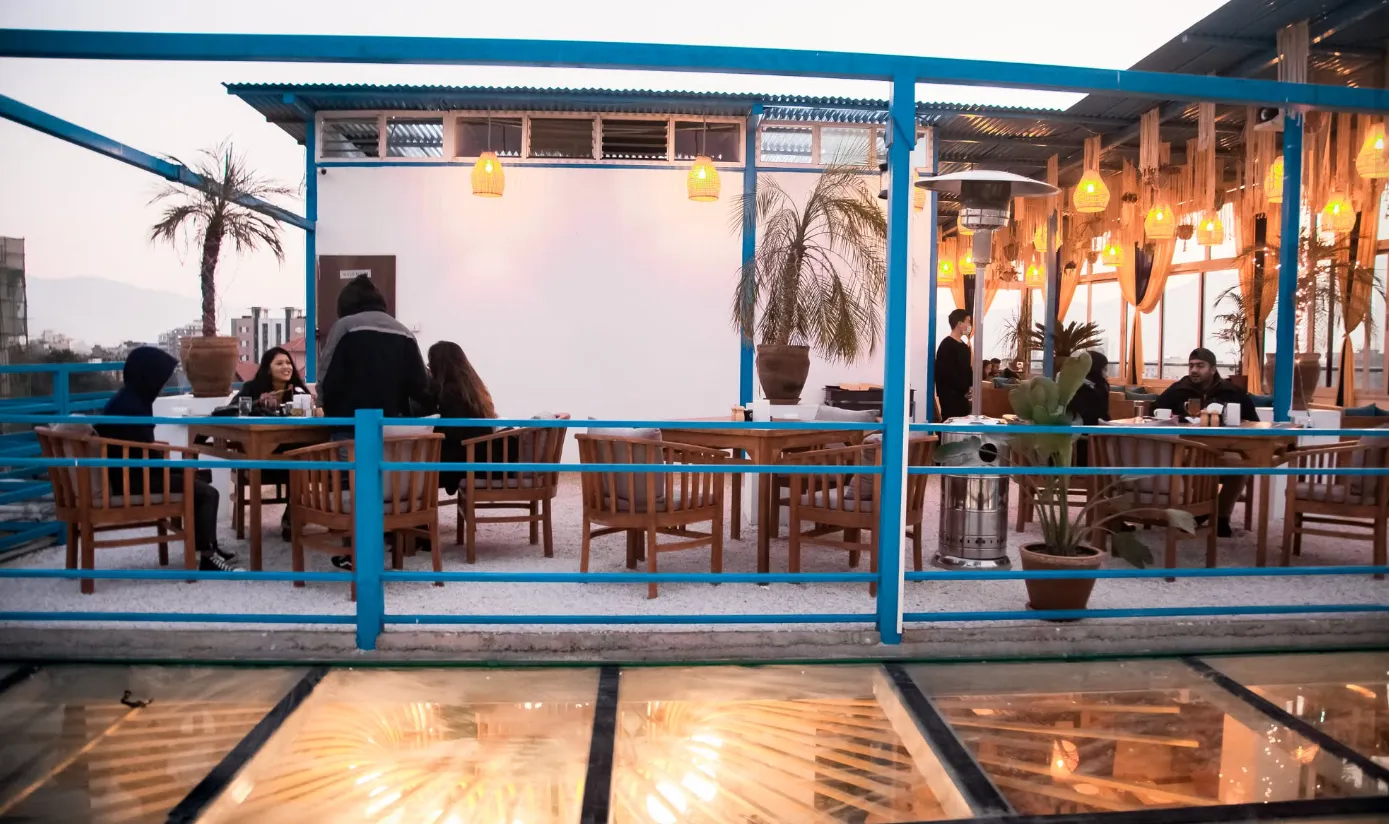 Rooftop dining tables with a lit glass floor and palm trees at dusk
